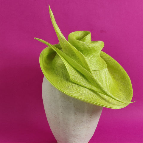 Lina Stein millinery workshops Ireland green sinamay fascinator with a swirled and curved design, displayed on a white mannequin head, set against a pink background.