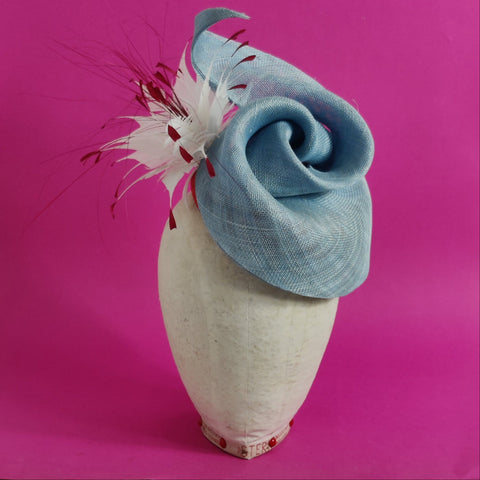 A light blue sinamay headpiece with a decorative element, displayed on a mannequin head.