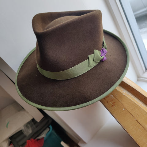Lina Stein millinery A brown felt trilby hat with a green brim, featuring a purple flower pin as a decoration.